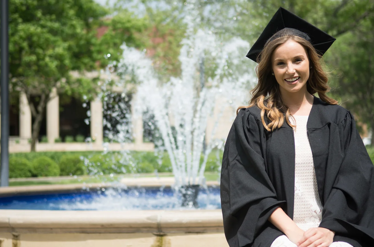 Emporia State student in graduation regalia in front of Plumb Hall fountain