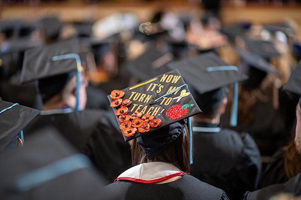 A group of students are seated in rows during the Emporia State University commencement ceremony, one wearing a hat that says "now it's my turn to teach!"