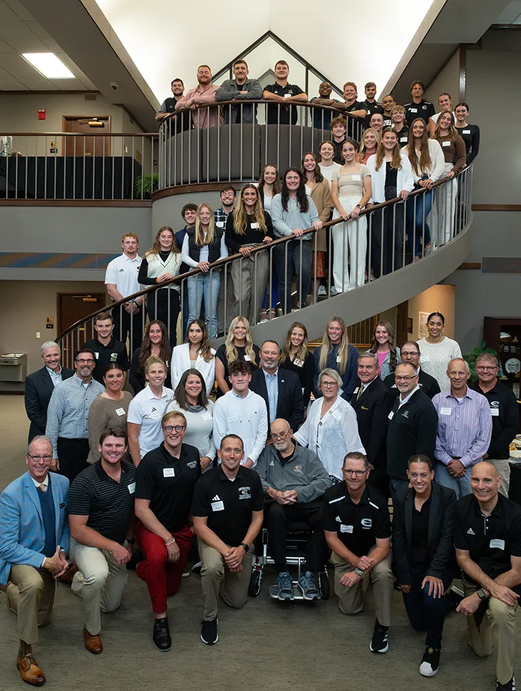 A group of 35 students standing on a spiral staircase with others in a group standing at the bottom of the stairs.